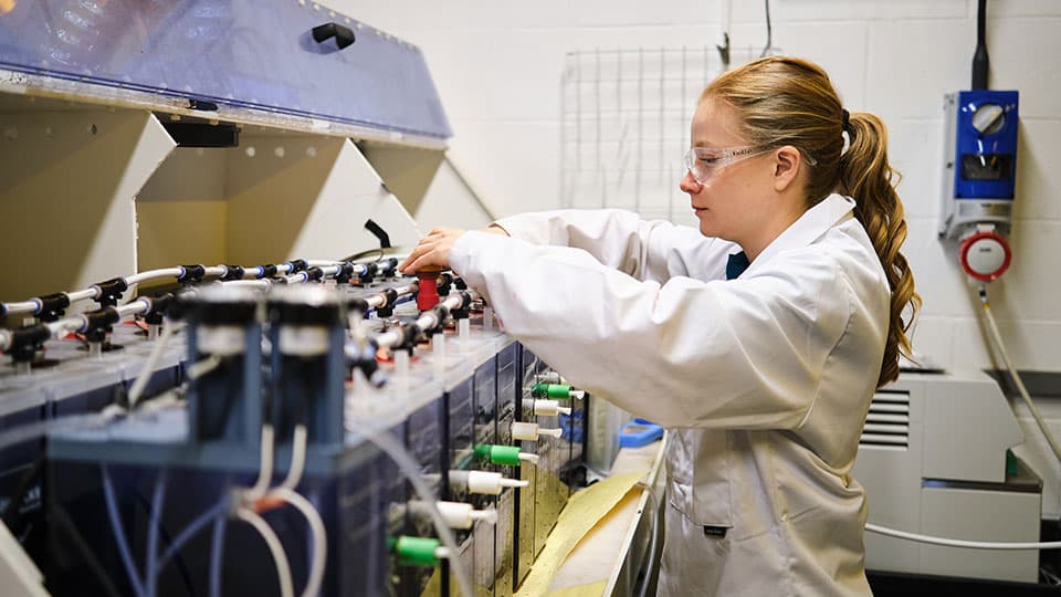 A woman in a lab coat and lab goggles positioning tubes on bottles